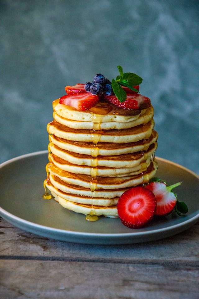 A stack of pancakes with blueberry & strawberries and poured honey presented on a grey plate.